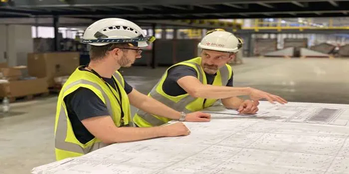 بهترین پیمانکار تاسیسات مکانیکی _ دو فرد با لباس کار که در حال گفتگو هستند و یک نقشه بزرگ جلو دست آنها قرار دارد.